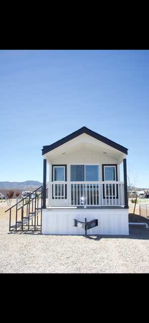 A small white manufactured home with a covered front porch and steps, set on a gravel lot under a clear blue sky.