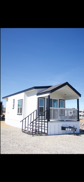A small white house with a black roof and railing, front porch, and steps, situated on a gravel lot under a clear blue sky.