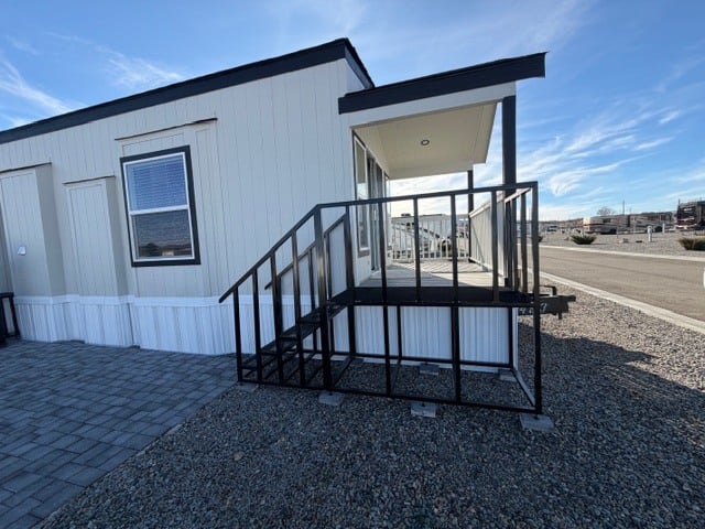 A modular building with a small porch, metal ramp railing, steps, and gravel ground in a paved, open parking area under a blue sky.