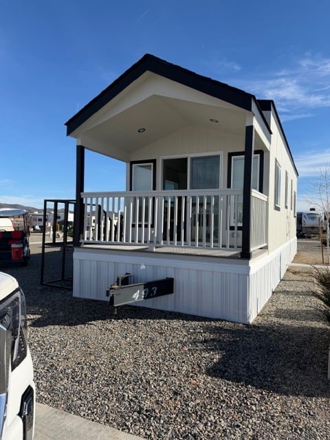 A small, modern manufactured home with a front porch and railing sits on a gravel lot under a clear blue sky.