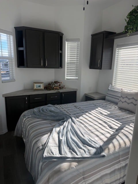 A neatly made bed with striped bedding is in a small bedroom with dark cabinets, a few windows, and sunlight streaming in. A blue-gray blanket is arranged in a heart shape on the bed.