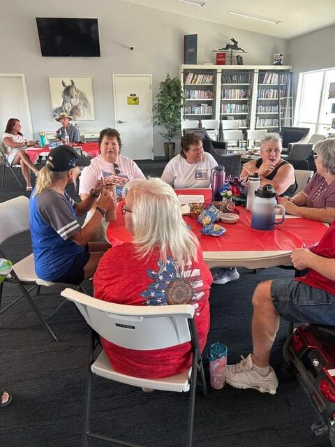 A group of people sit around a table with red coverings, chatting and eating snacks in a room with bookshelves and wall art in the background.