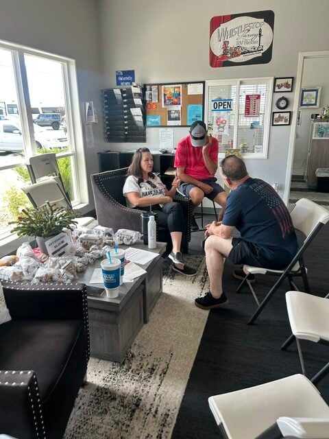 Three people sit and talk in a waiting area with snacks, drinks, and informational items on a table. Sunlight comes through large windows. A bulletin board and office decor are visible on the wall.