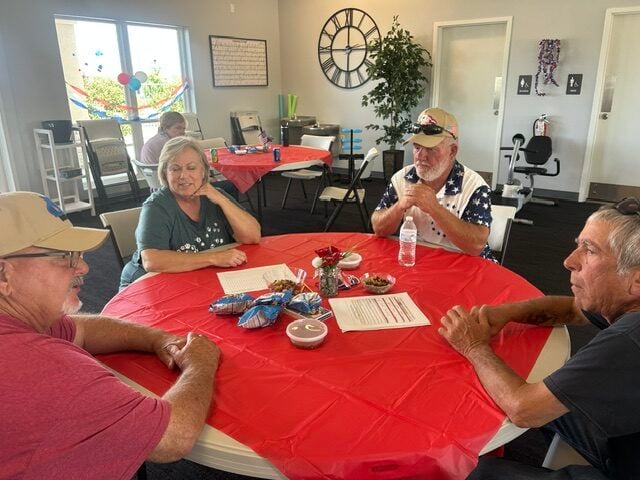 Four older adults sit around a red tablecloth, talking and sharing snacks in a bright, casual room with decorations and large wall clock.