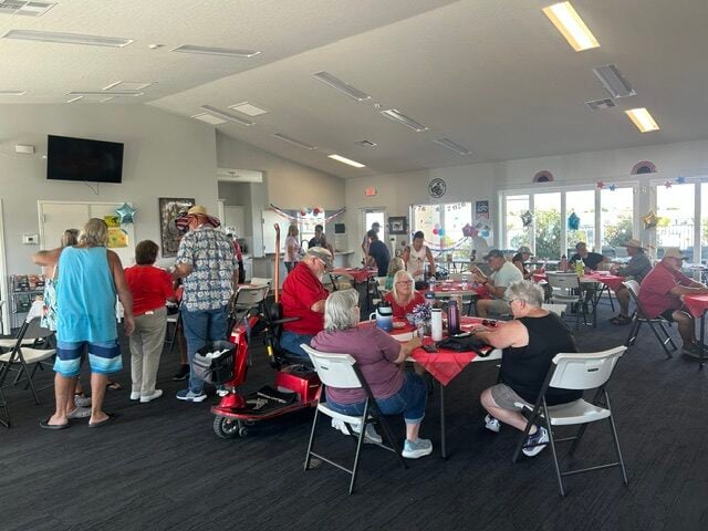 People are gathered in a community room, sitting at tables and socializing. Some decorations are visible, and the atmosphere appears casual and friendly.