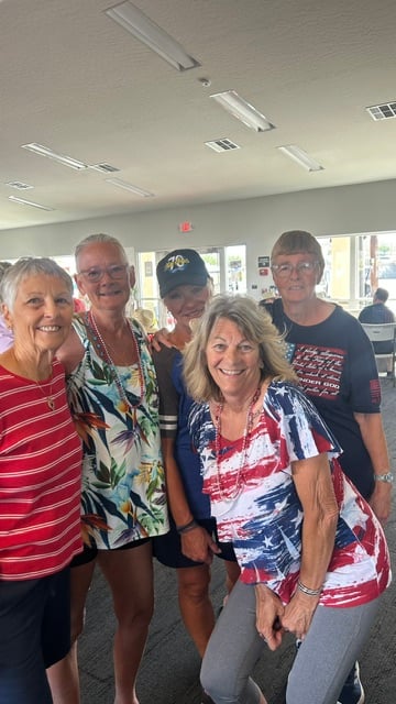 Five older women stand together indoors, smiling at the camera. They wear casual summer clothing, some with patriotic or tropical patterns. The setting appears to be a community or event space.